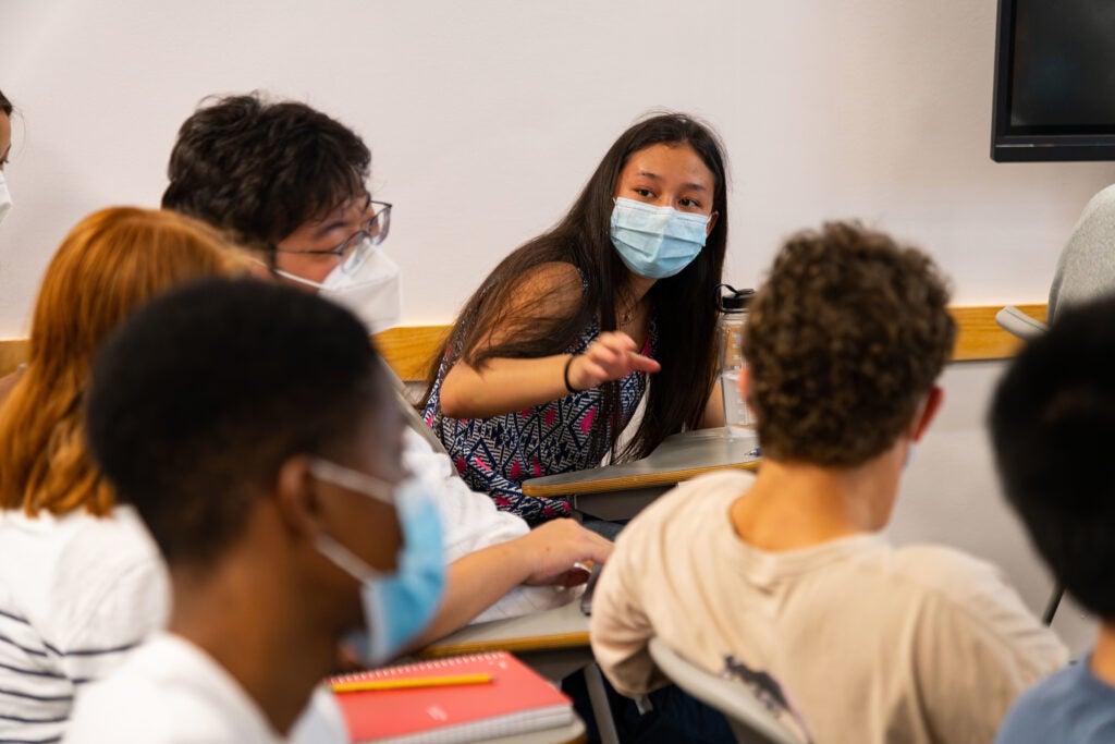 Young woman in a mask speaks to classmates sitting at desks around her