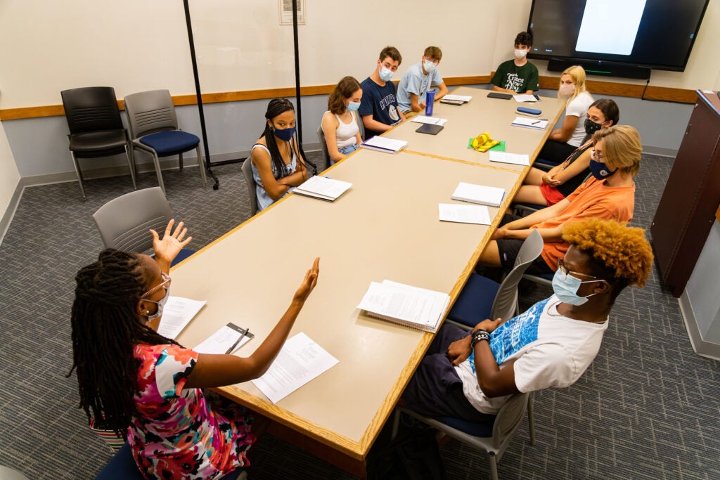 Students sit around a table with papers and tables while watching a teacher at the end of the table speak with her hands.