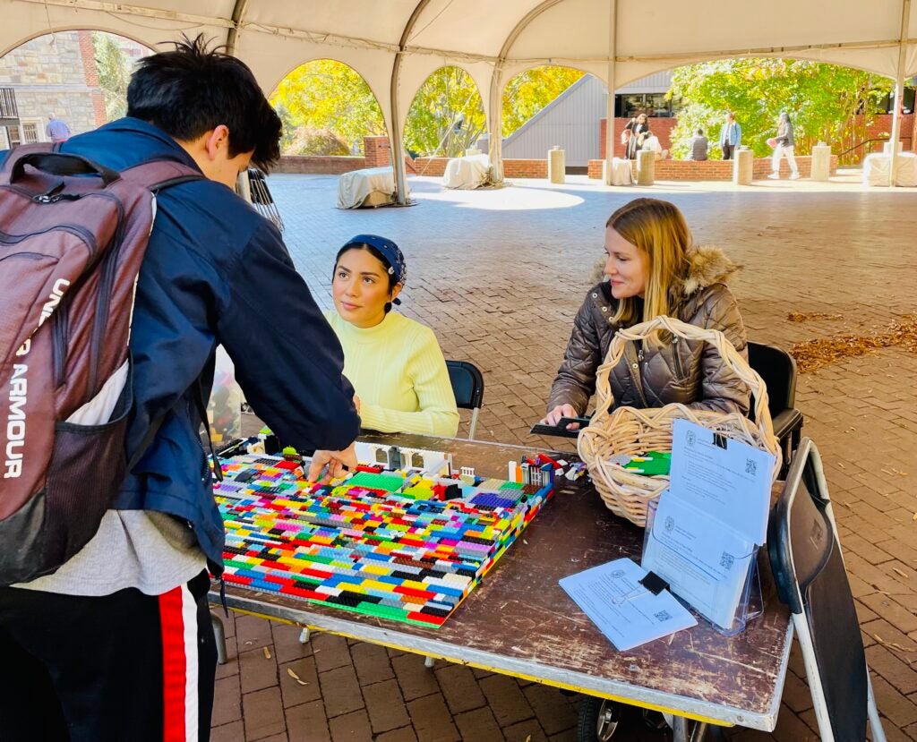 Young man leans over a lego ramp while two women watch seated on the other side of the table outside