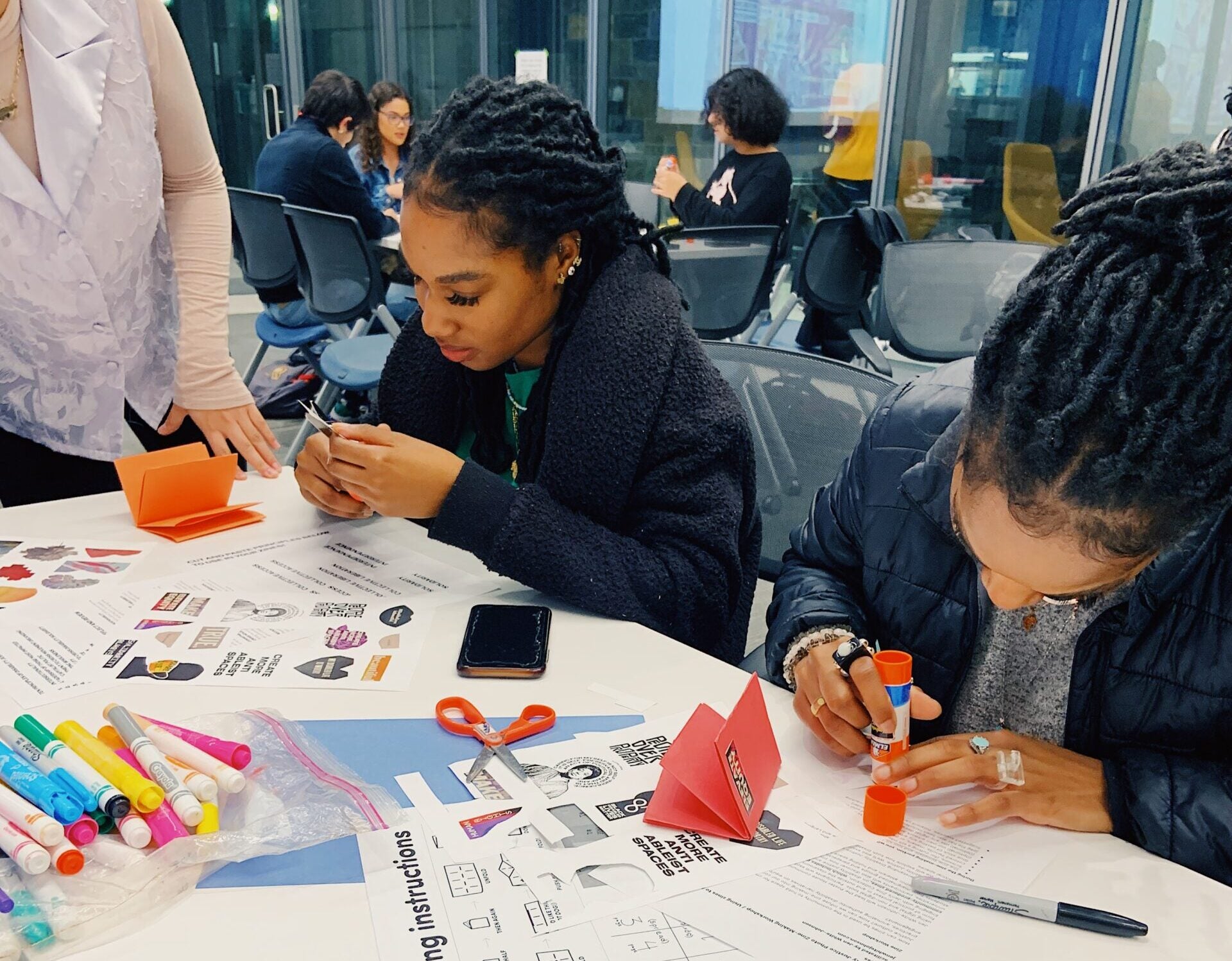 Young women make zines with paper, markers, glue and scissors spread out in front of them.
